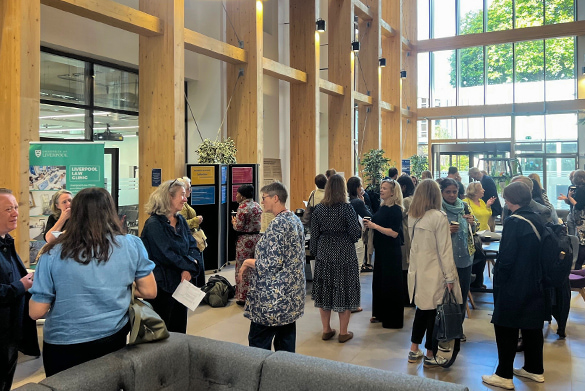 Group of people conversing in a modern lobby with wooden beams and tall windows; a green sign reading \'Liverpool Law Clinic\' is visible on the left