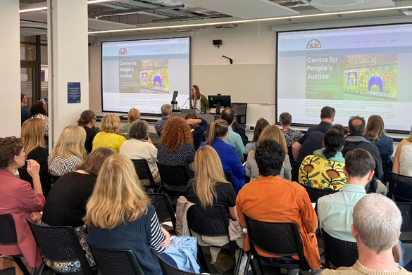 Audience seated in a room, listening to a speaker at a podium with two screens behind displaying 'Centre for People's Justice'.