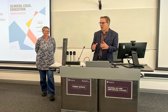 Two individuals stand at the front of a lecture hall at the University of Liverpool's School of Law and Social Justice. One person stands to the left, while the other speaks from behind a podium.