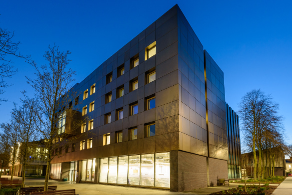 The School of Law and Social Justice. A modern multi-story building with a sleek, dark metallic facade and rows of square windows, illuminated from inside at dusk.