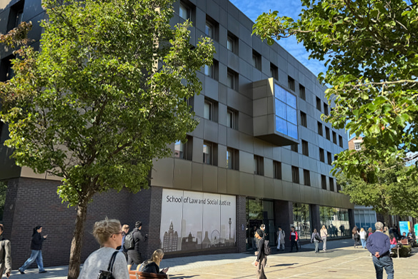 A modern building with a sign reading \'School of Law and Social Justice\'. Several people are walking and sitting outside, with trees lining the sidewalk under a clear blue sky.