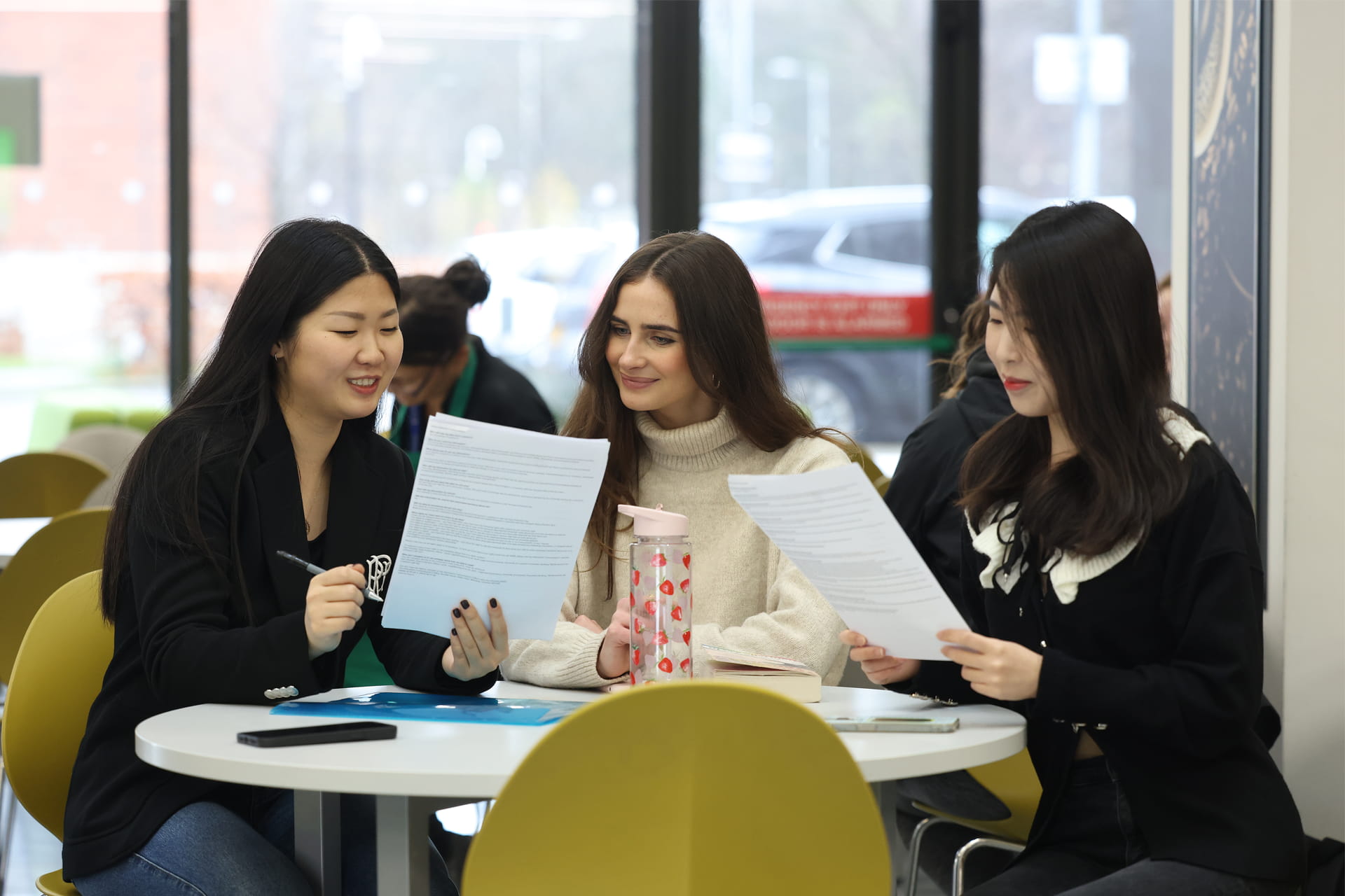 Three students seated at a round table indoors, reviewing printed documents.