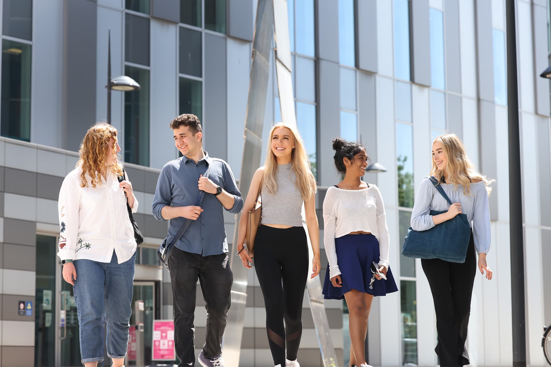 Five people walk together outside a modern building with large windows and vertical panels, carrying bags and conversing.