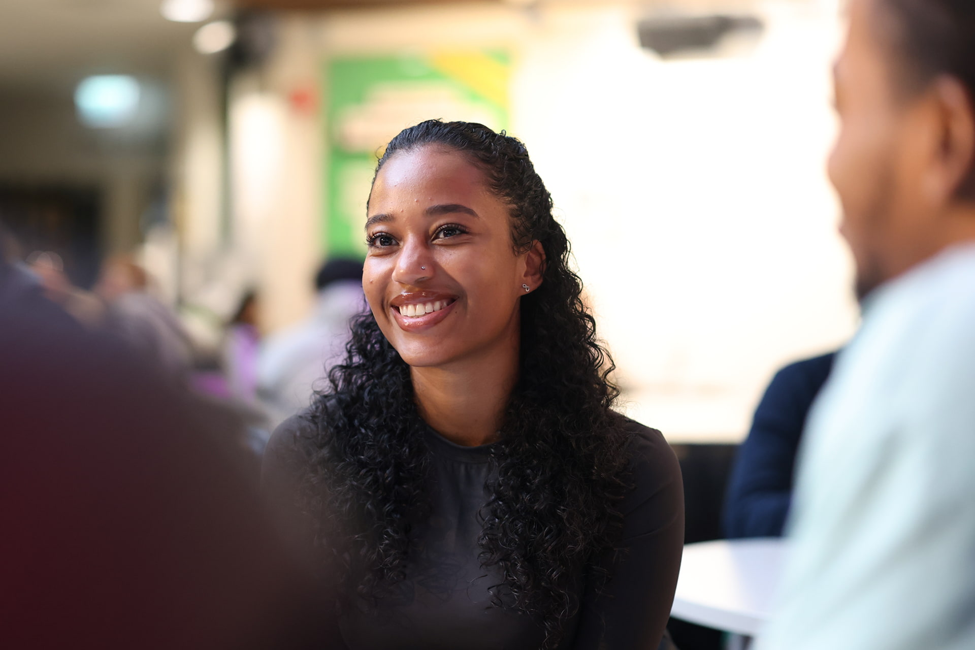 A student sits indoors in a well-lit public space. The background includes blurred figures.