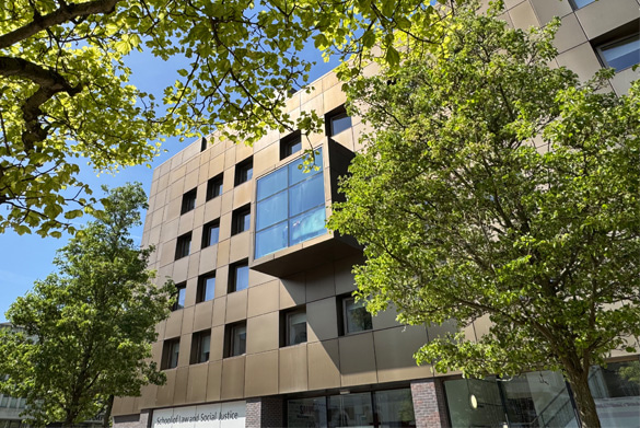 The exterior of the School of Law and Social Justice Building, featuring a modern bronze façade with large windows, framed by several leafy trees in the foreground.