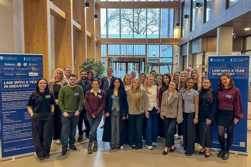 A group of placement students and industry partners standing together in a bright, modern atrium beside two large blue display boards titled ‘Law with a Year in Industry\'.