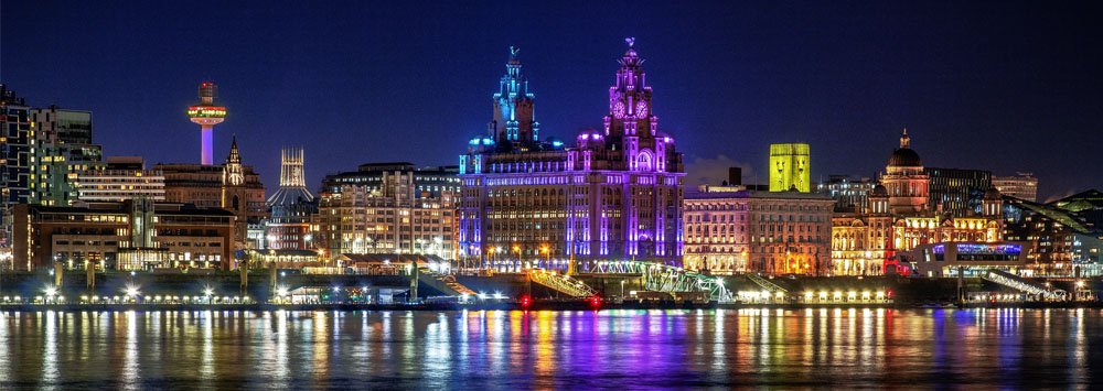 Liverpool skyline at night. The skyline is reflected in the river. The Liver Bird is lit up with purple and blue lights.