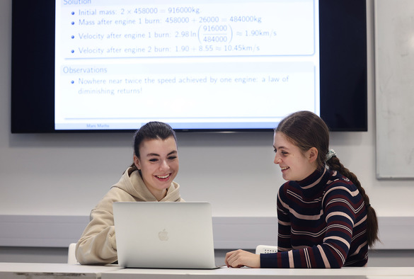 Two students collaborate at a table, focused on a laptop in front of them.