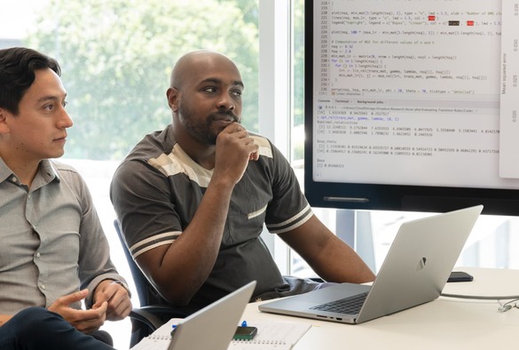 Two men seated at a table, working on laptops with a computer screen displaying information between them