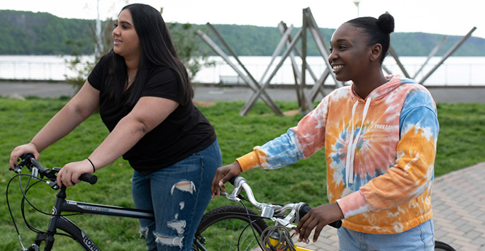 Two young women pushing their bikes in a green space