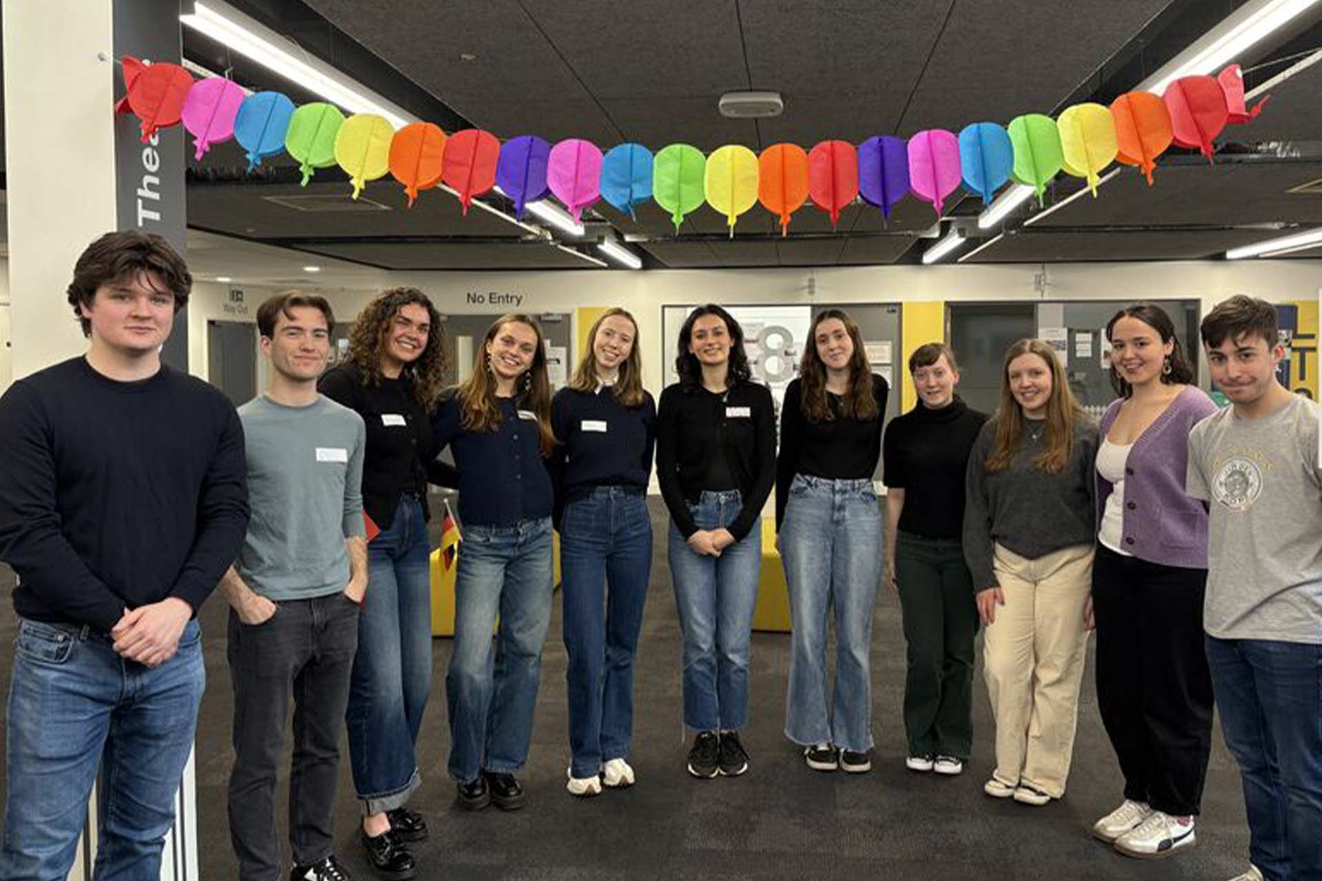 A group of students smiling and standing together under colourful bunting at an event.