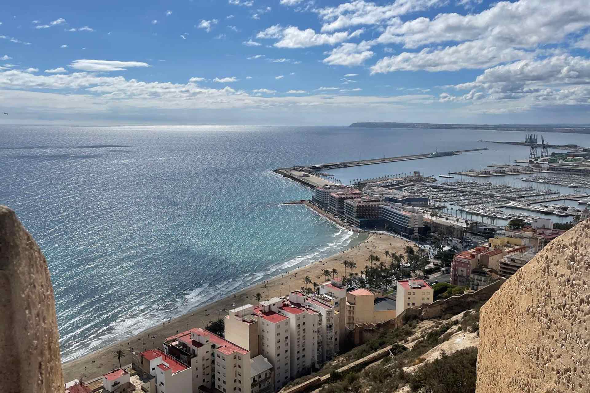 A top down view of a sunny European coastline with a sandy beach, in the foreground are stone walls and in the distance is a harbour and white apartment blocks.