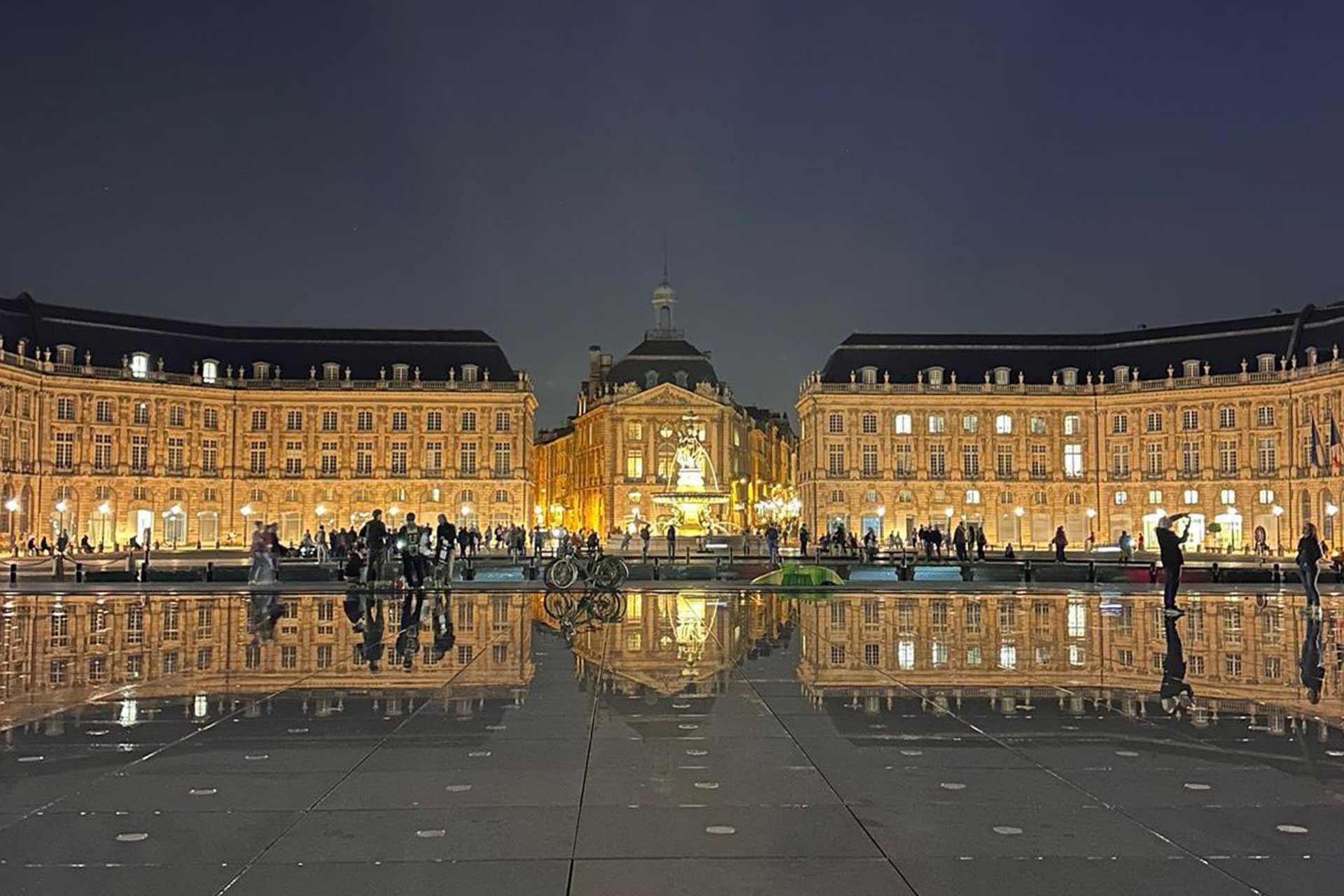 A grand city centre square at night with old French buildings lit up and a statue in the centre. Tourists take photos on the square.