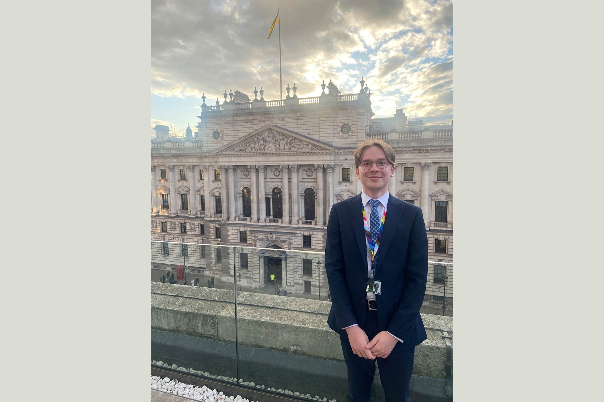 A young man standing and smiling posing in front of a parliamentary building.