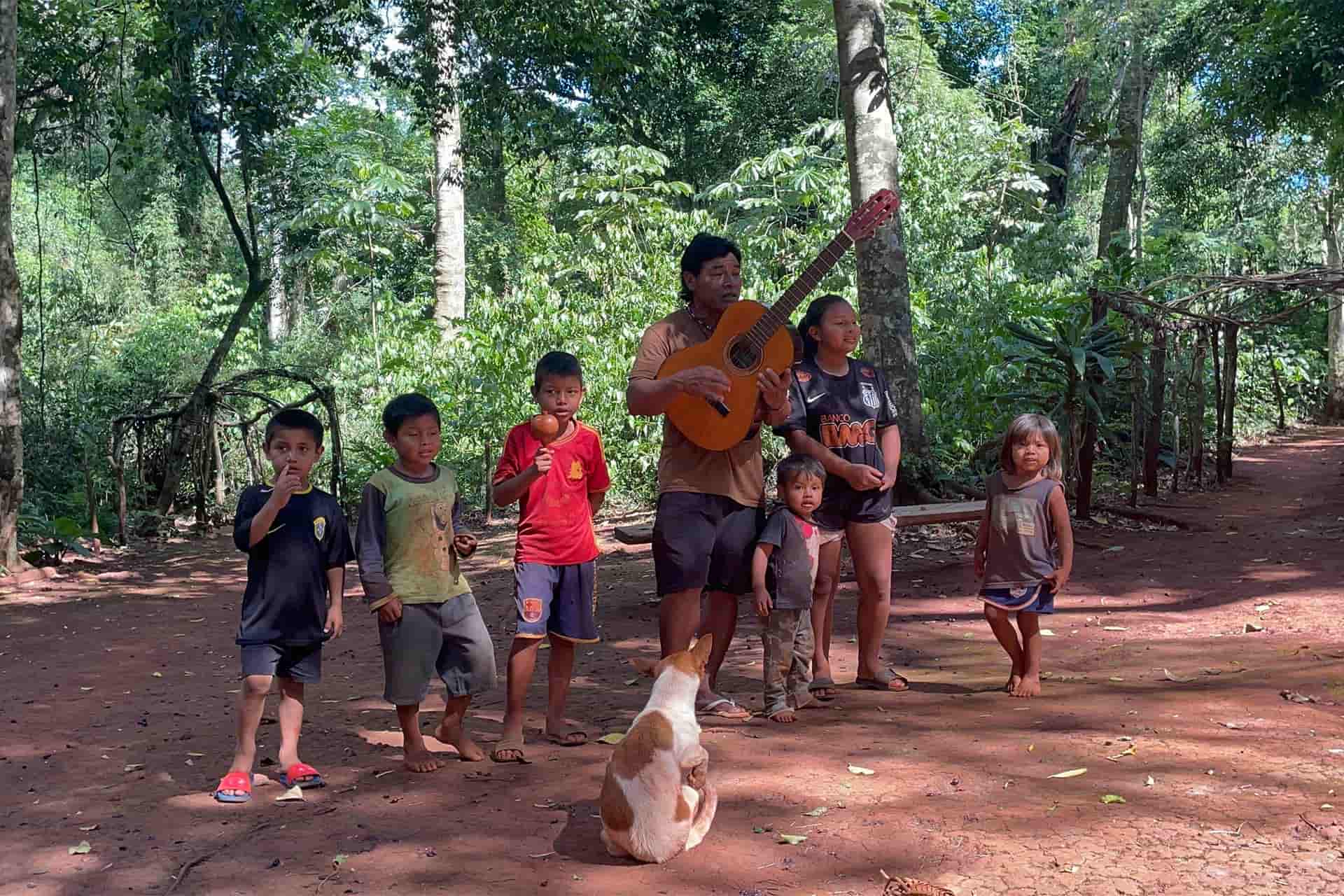 5 children singing and an adult playing a guitar in a wooded area.