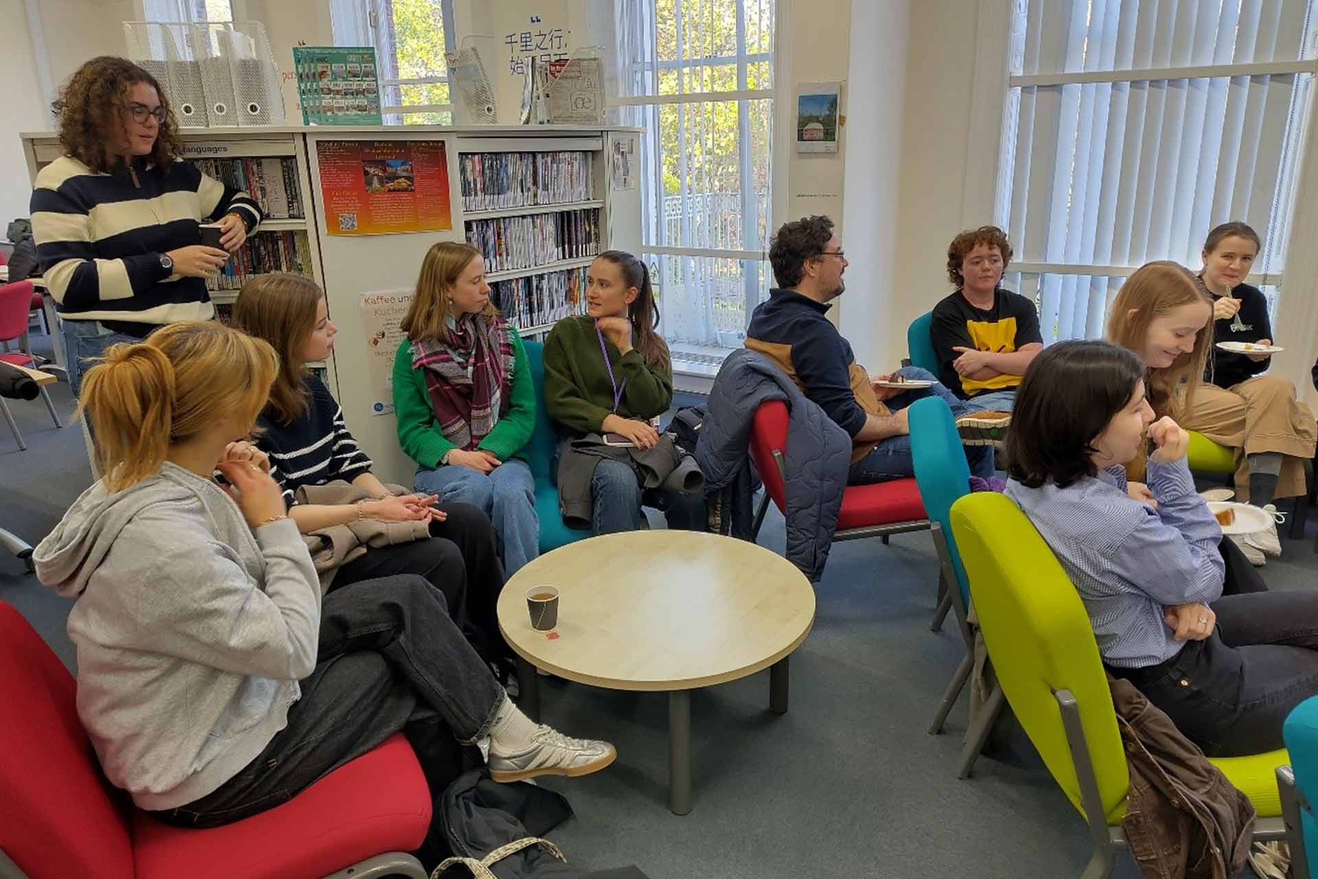 A group of students sitting in a lounge with bookcases, talking and eating cake and drinking coffee