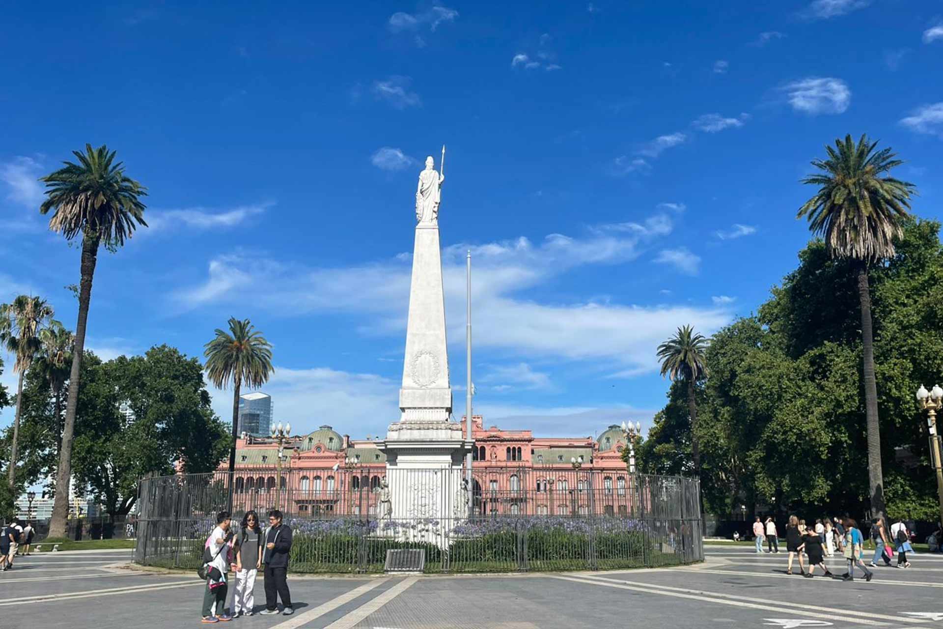 A city square with palm trees with a tall white statue in the centre.