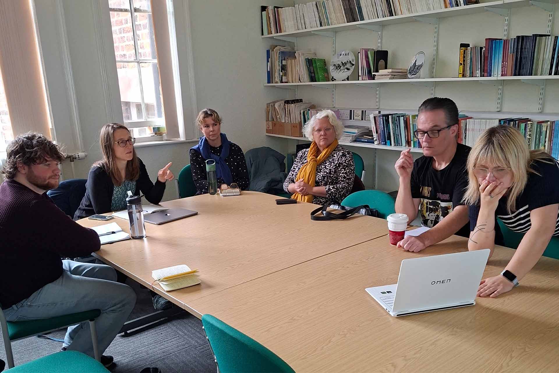 Four women and two men in discussion, sitting around a table looking at a laptop screen.