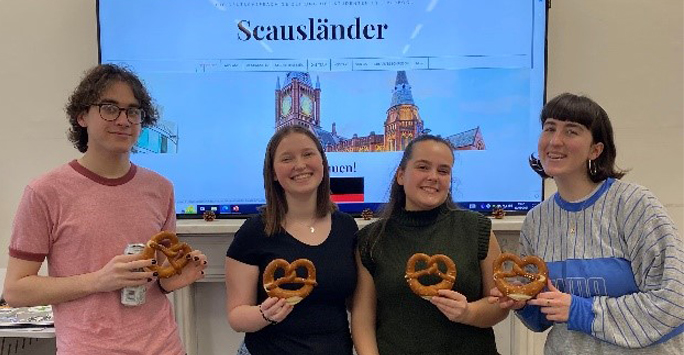 4 students smiling at the camera holding pretzels in front of a screen showing a website displaying Scauslander student newspaper