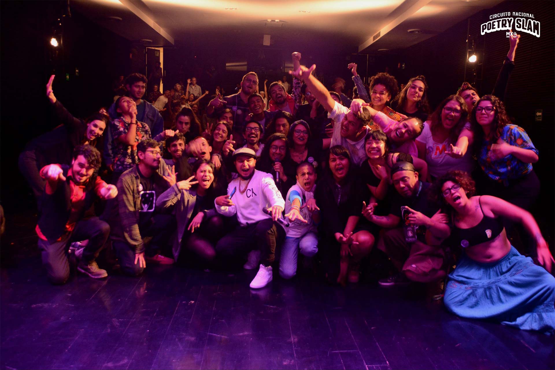 A group of people inside a room, a person in the centre holding a microphone whilst others smile and laugh with their arms and hands in the air.