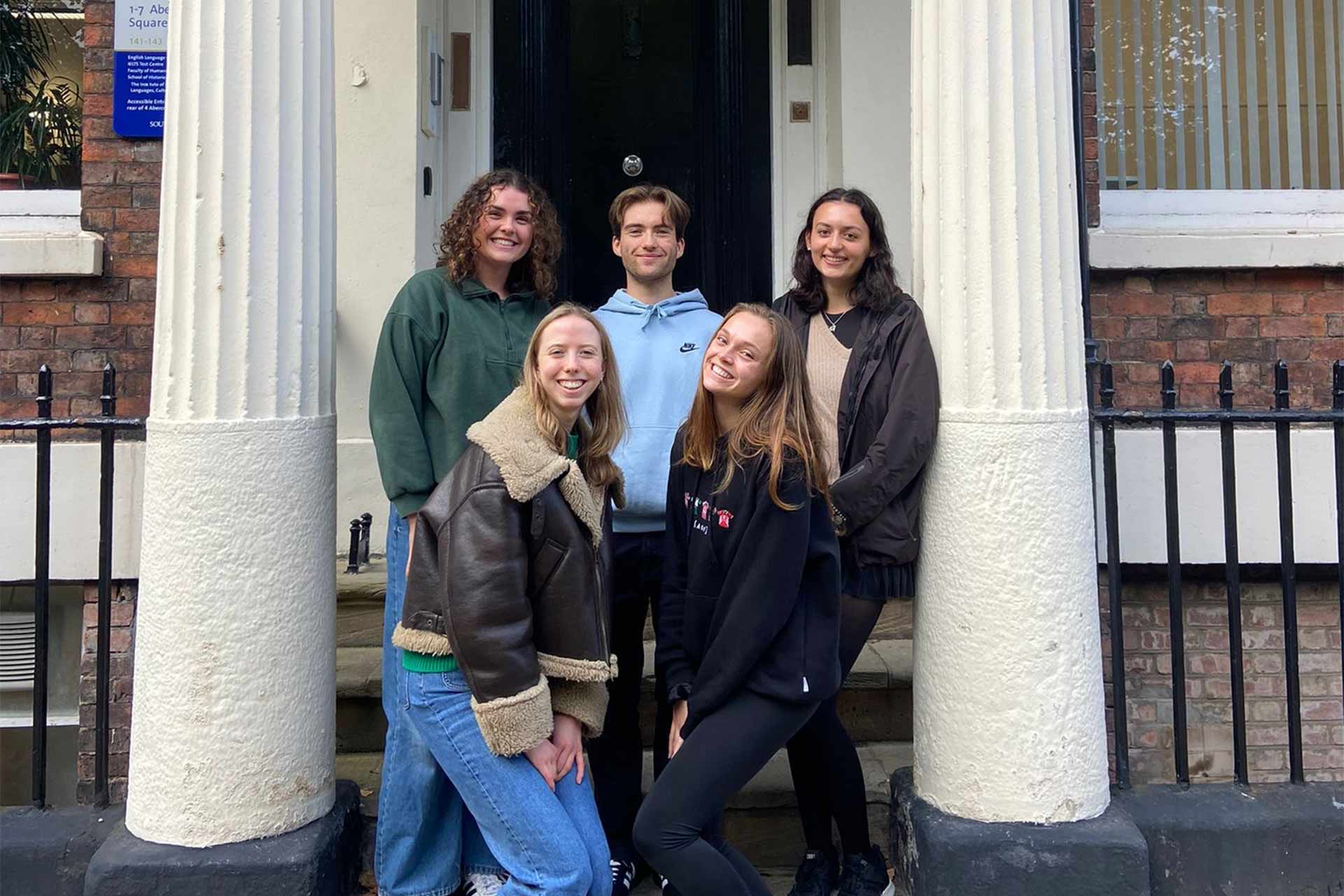 5 students, comprising of 1 man and 4 women standing together and smiling outside a university campus building.