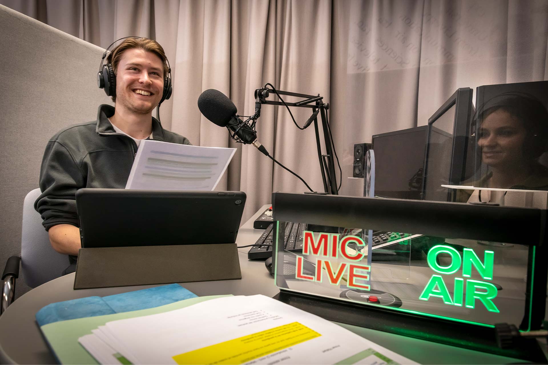 A student with a microphone and headphones on smiling and reading a piece of paper with a lit up sign in front of him reading 'MIC LIVE' and 'ON AIR'
