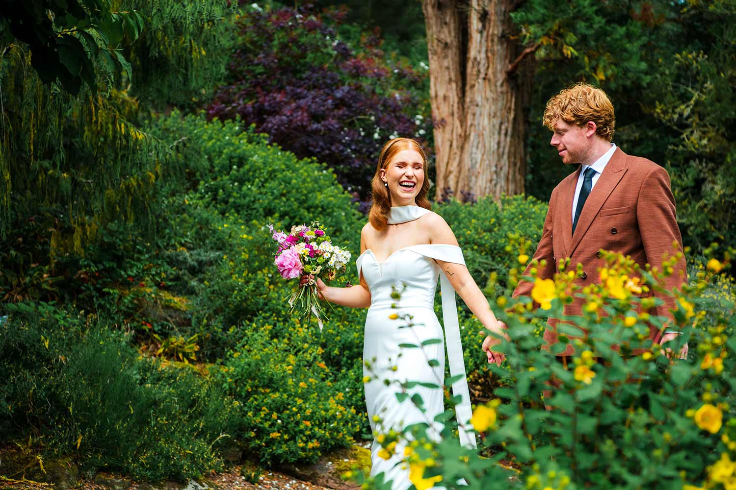 A couple walk through the gardens after their wedding, the bride holding a bouquet.