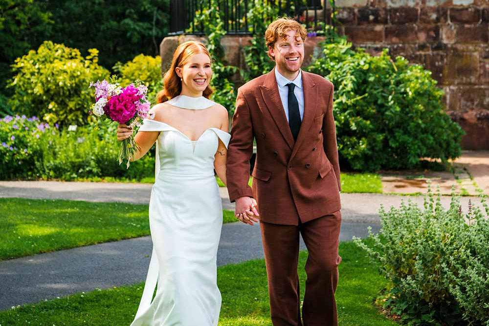 A couple walk through the gardens at Ness, the bride holding a bouquet, on the way to their wedding reception.