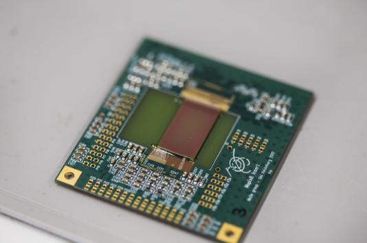 Photograph of the MuPix8 prototype for the Mu3e experiment mounted on its carrier board [photo credit Niklaus Berger]. This experiment will search for the decay of a muon to three electrons. Any measurement of this decay would be a clear sign of new physics. The Mu3e requires tracking detectors with very low mass (50 μm thin) and excellent timing resolution (