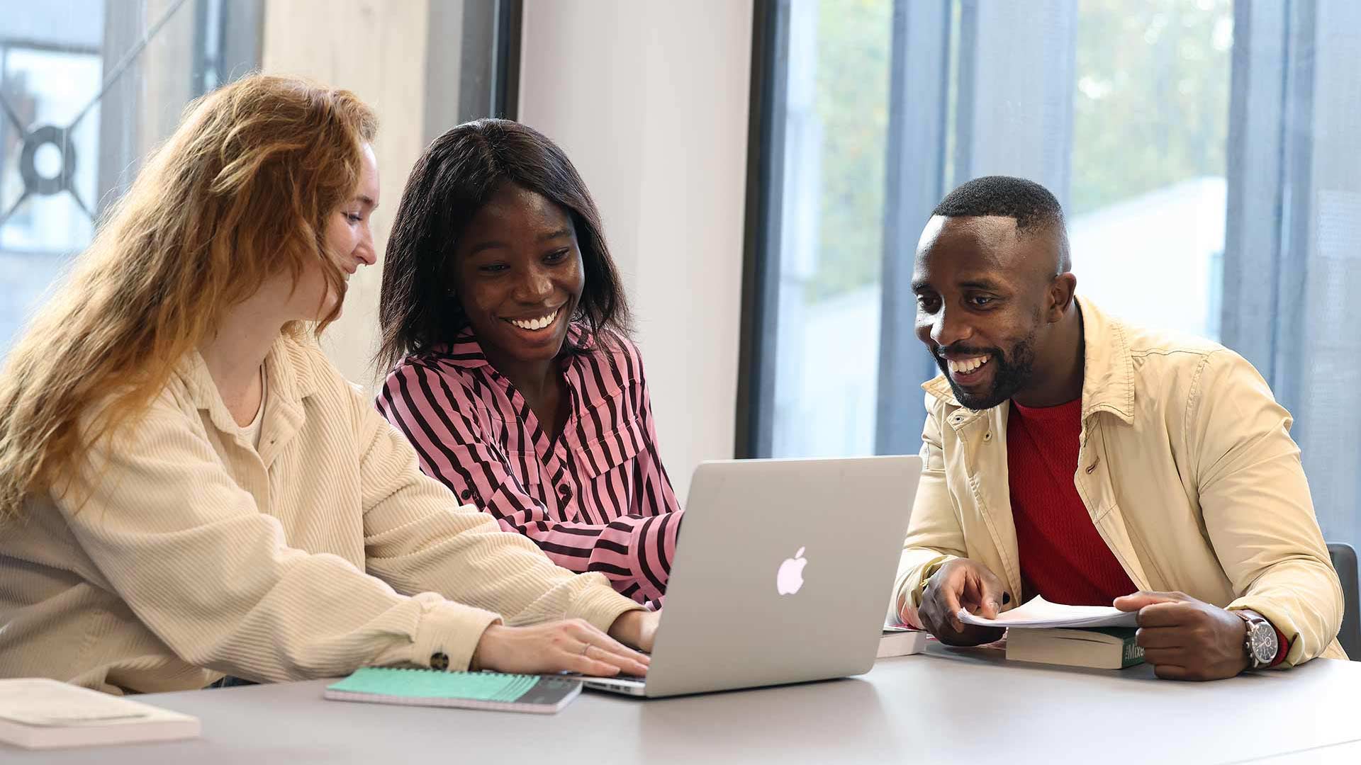 Three students in a classroom looking at a Macbook screen.