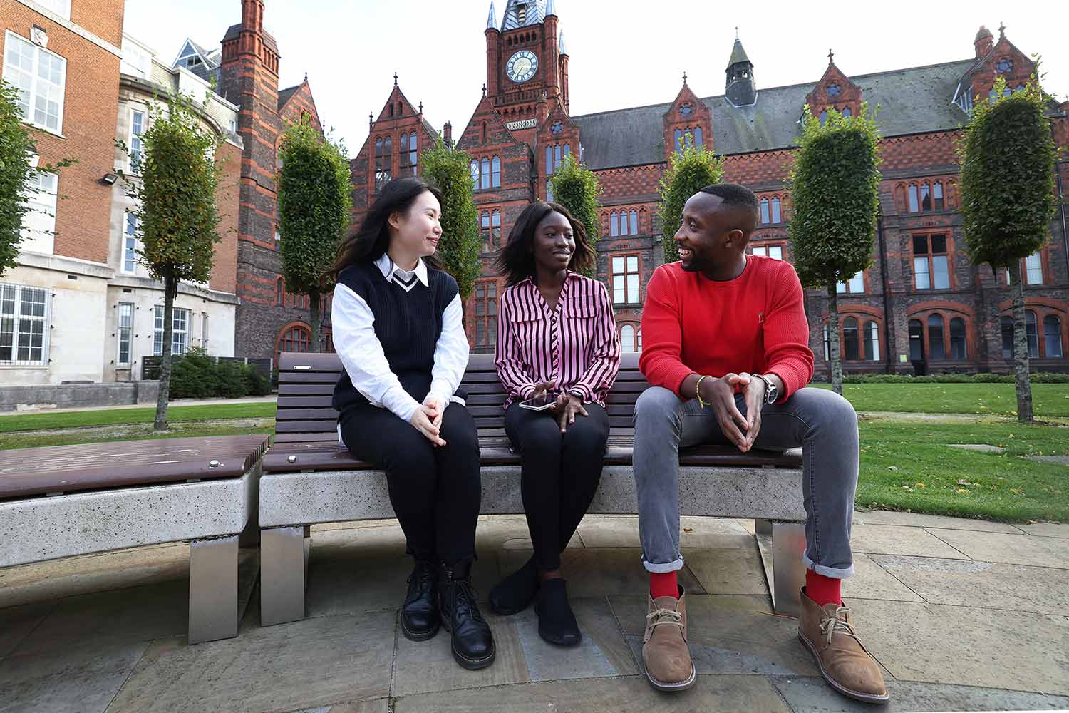 Three students sitting on a bench in the Quad with the Victoria Gallery & Museum visible behind them.