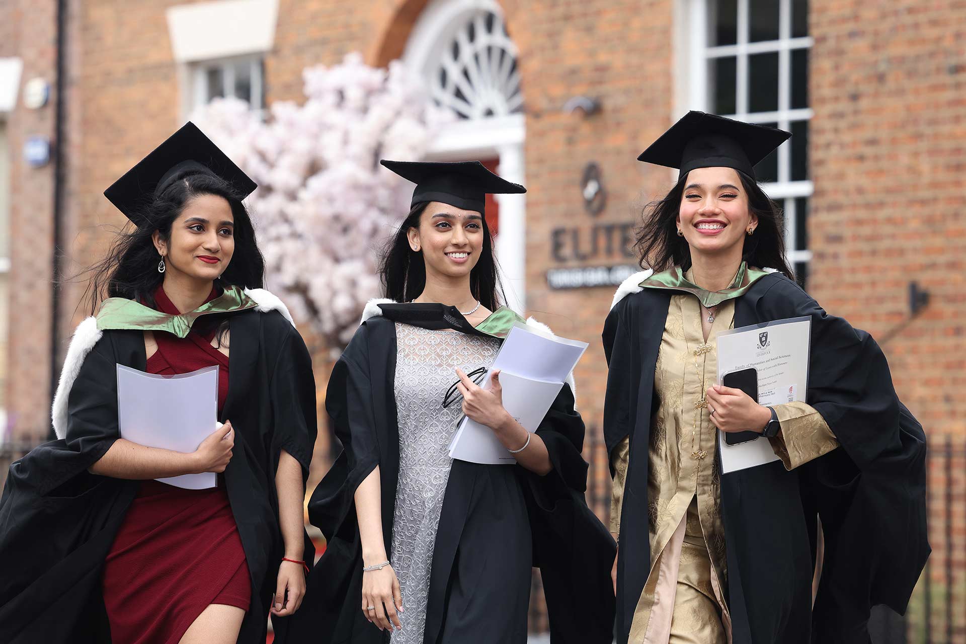 Three graduates in their cap and gowns walking on campus, carrying their degree certificates.