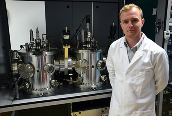 John Major in a lab coat stands beside various machines in a laboratory setting, showcasing a scientific environment.