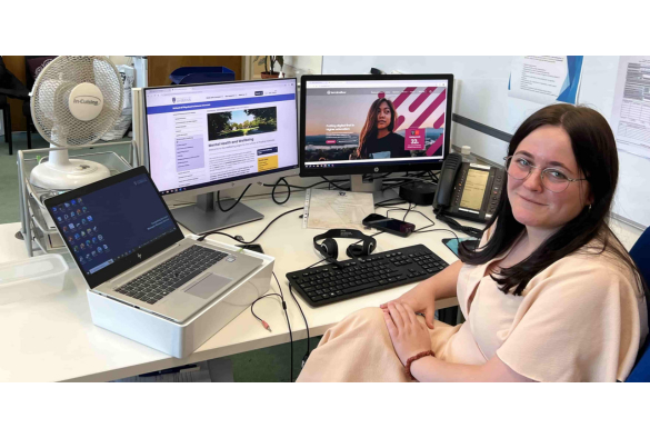 Hannah Melia sits at a desk with two computers and a fan