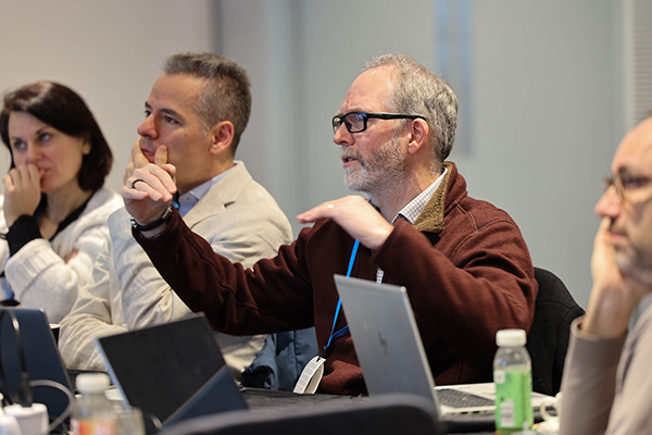 People sitting at a desk with laptops in front of them. One person talking.