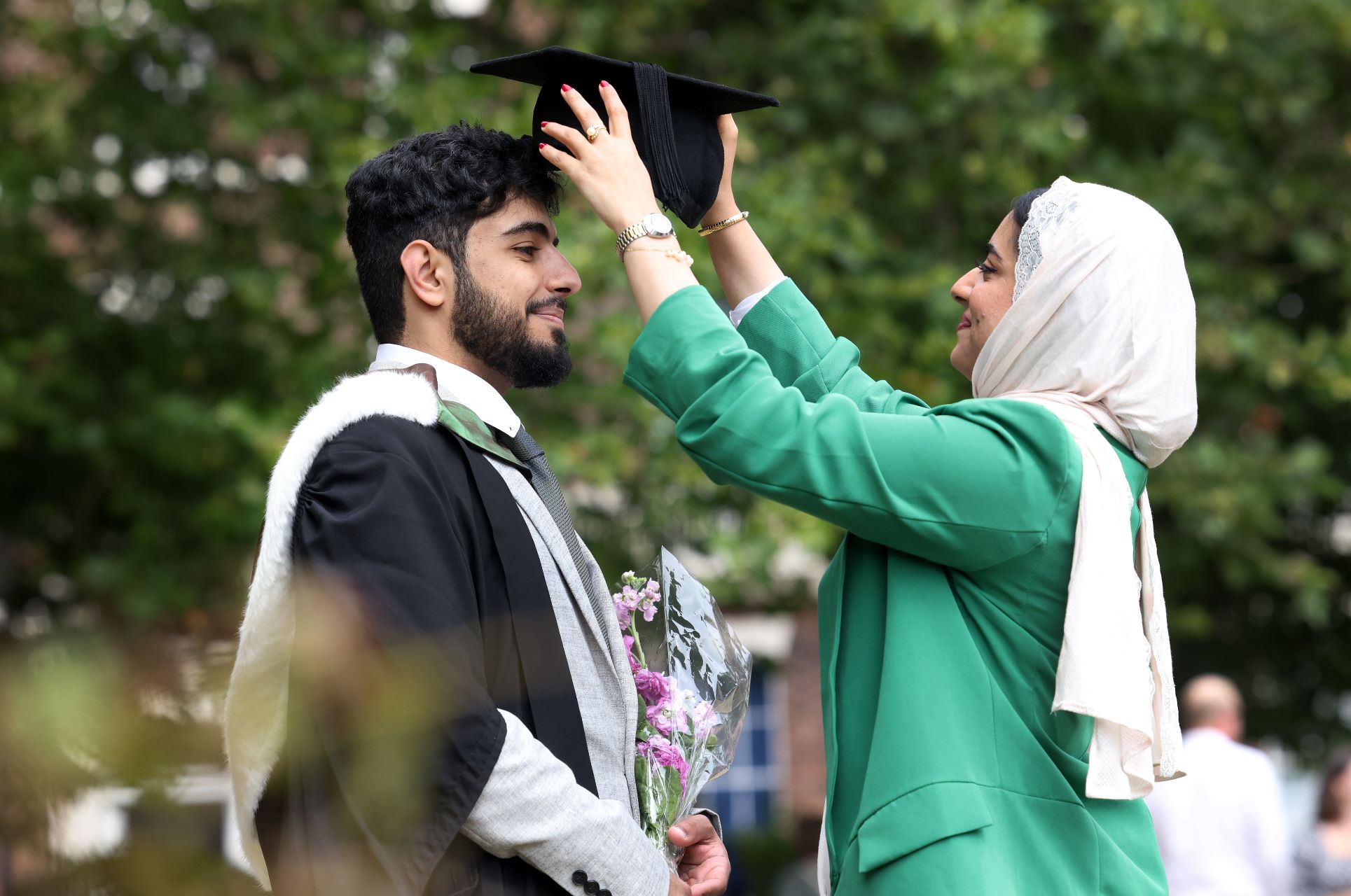 Two people at graduation. One person, wearing green, is placing a graduation cap on the head of a student dressed in graduation robes.