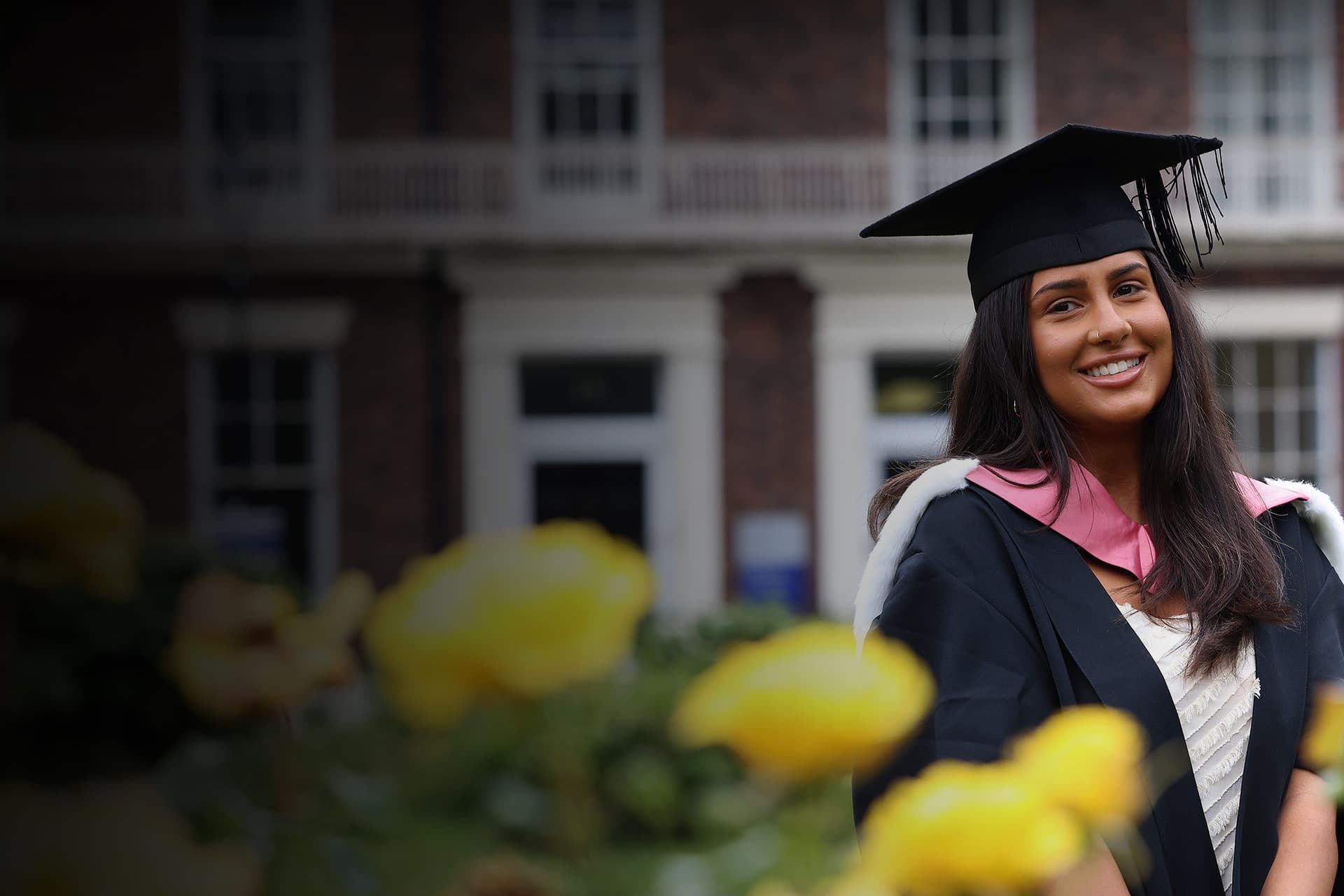 A student dressed in graduation robes standing behind yellow flowers.