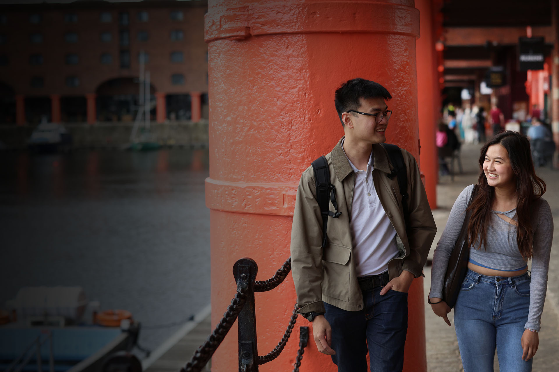 Two people in front of a red pillar at the docks in Liverpool, smiling.
