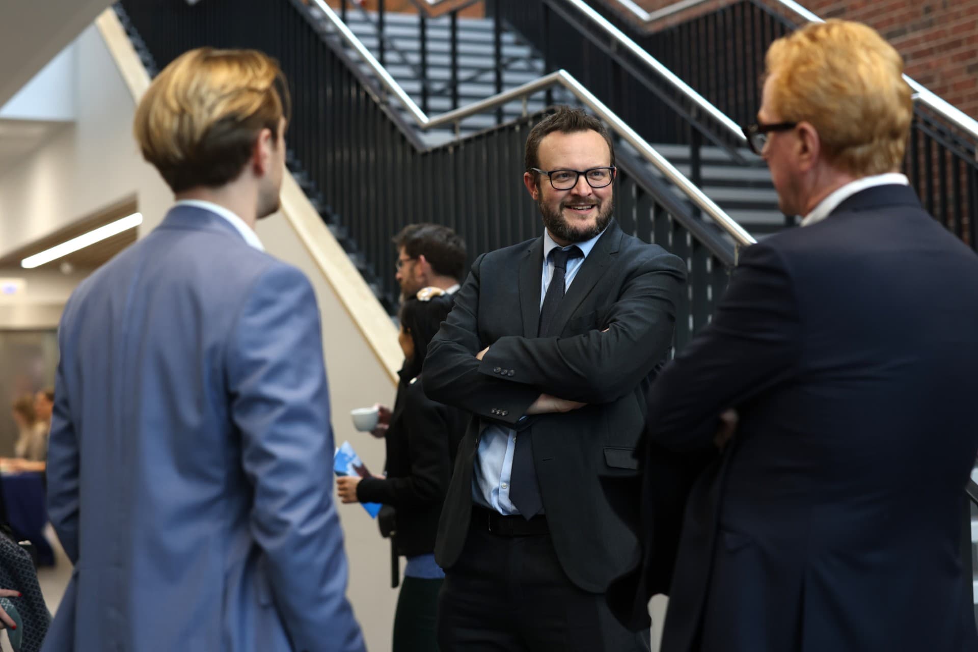 Three people standing talking wearing suits. One of the people is facing the camera and smiling, with the other two people facing away.