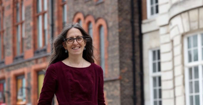 Georgina Engfield is smiling and walking on campus. She is wearing a red dress, glasses, and has dark hair.