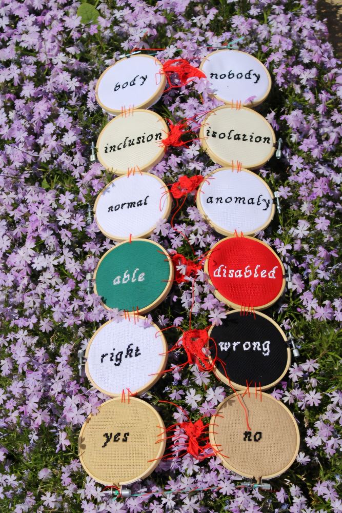 Hand-embroidered textile artwork made from embroidery hoops and cross-stitch fabric, featuring paired words—body/nobody, inclusion/isolation, normal/anomaly, able/disabled, right/wrong, yes/no—interconnected by the word “binary” crocheted in red thread through the centre. Mixed textile, approximately 80×30 cm.