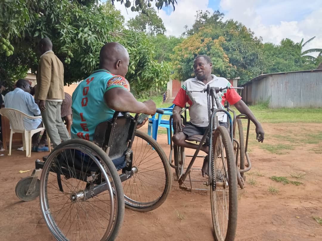 Two African men in wheelchairs are engaged in conversation on a compound with a concrete wall and a grassy surface. The man facing towards the camera has a clear disability affecting his legs. Behind the men is a small group of individuals sitting under the shade of a mango tree.