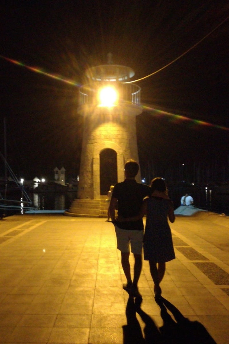 Two people walk side by side along a seaside path at night, their backs to the camera. A lighthouse’s beam lights the dark surroundings, symbolising guidance and companionship.