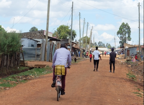 A man cycling down a dirt road with a yellow package on his bike. In the background are other people walking on the road