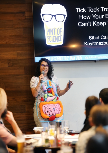 Sibel wearing an apron with organs on wearing wristwatches. Pint of Science logo is seen on the screen at the background.