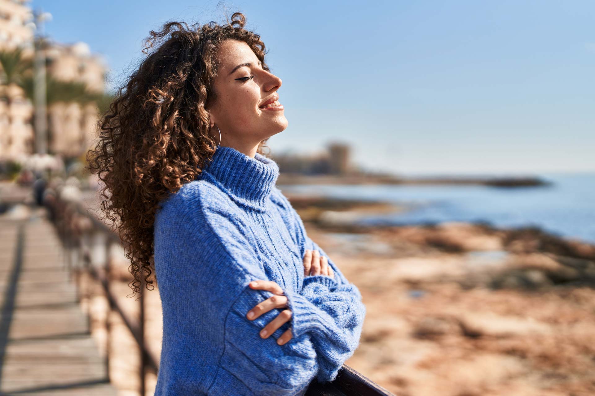 A woman is standing in a coastal area with her eyes closed and arms crossed. She looks as if she is breathing in the clean air.