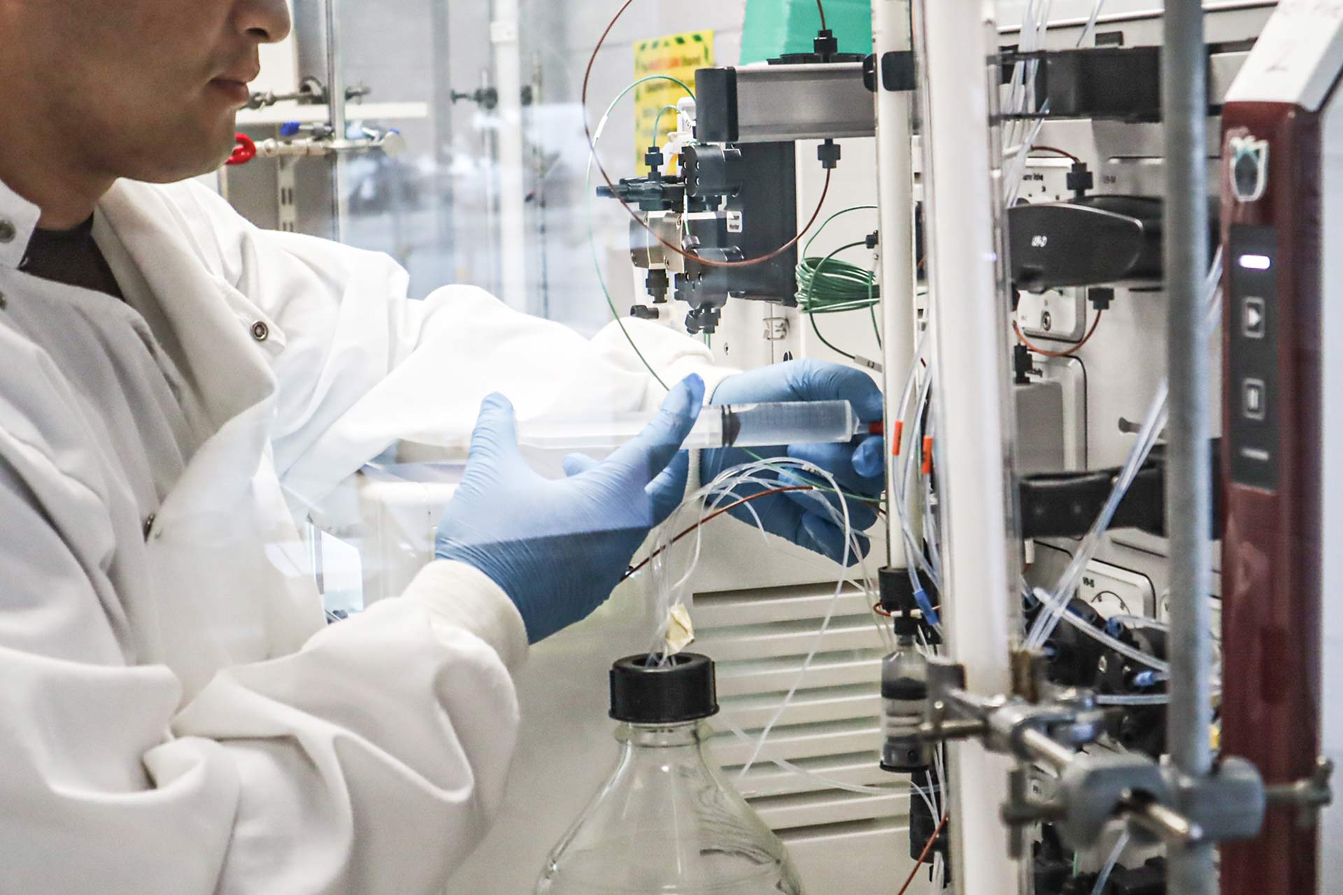 A person in a lab coat and blue gloves adjusts wires and tubing on laboratory equipment next to a large glass bottle.