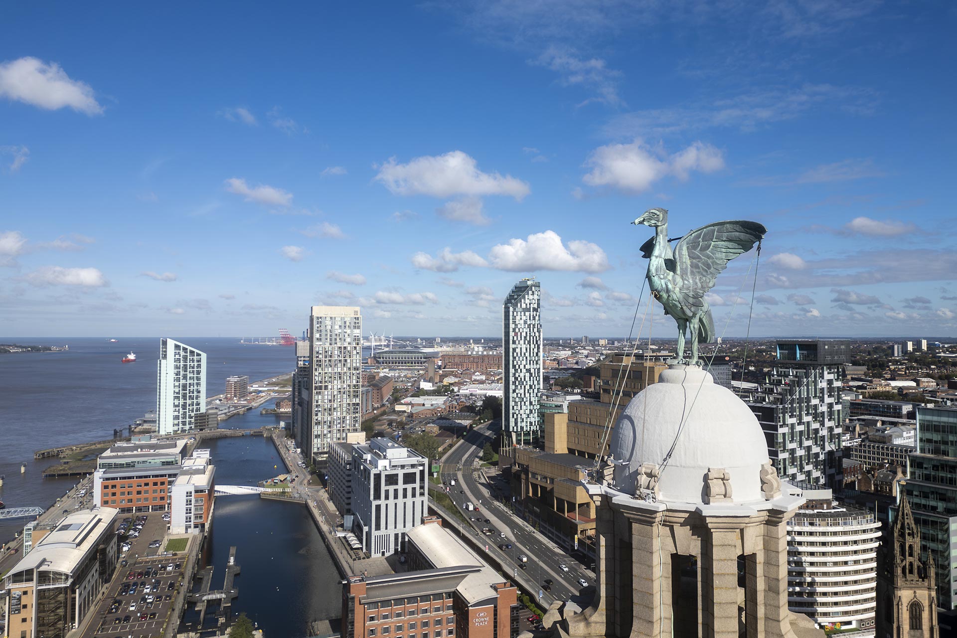 Aerial photograph of Liverpool's skyline featuring the River Mersey and various buildings on the waterfront. One of the Liver Bird statues is prominent in the foreground.