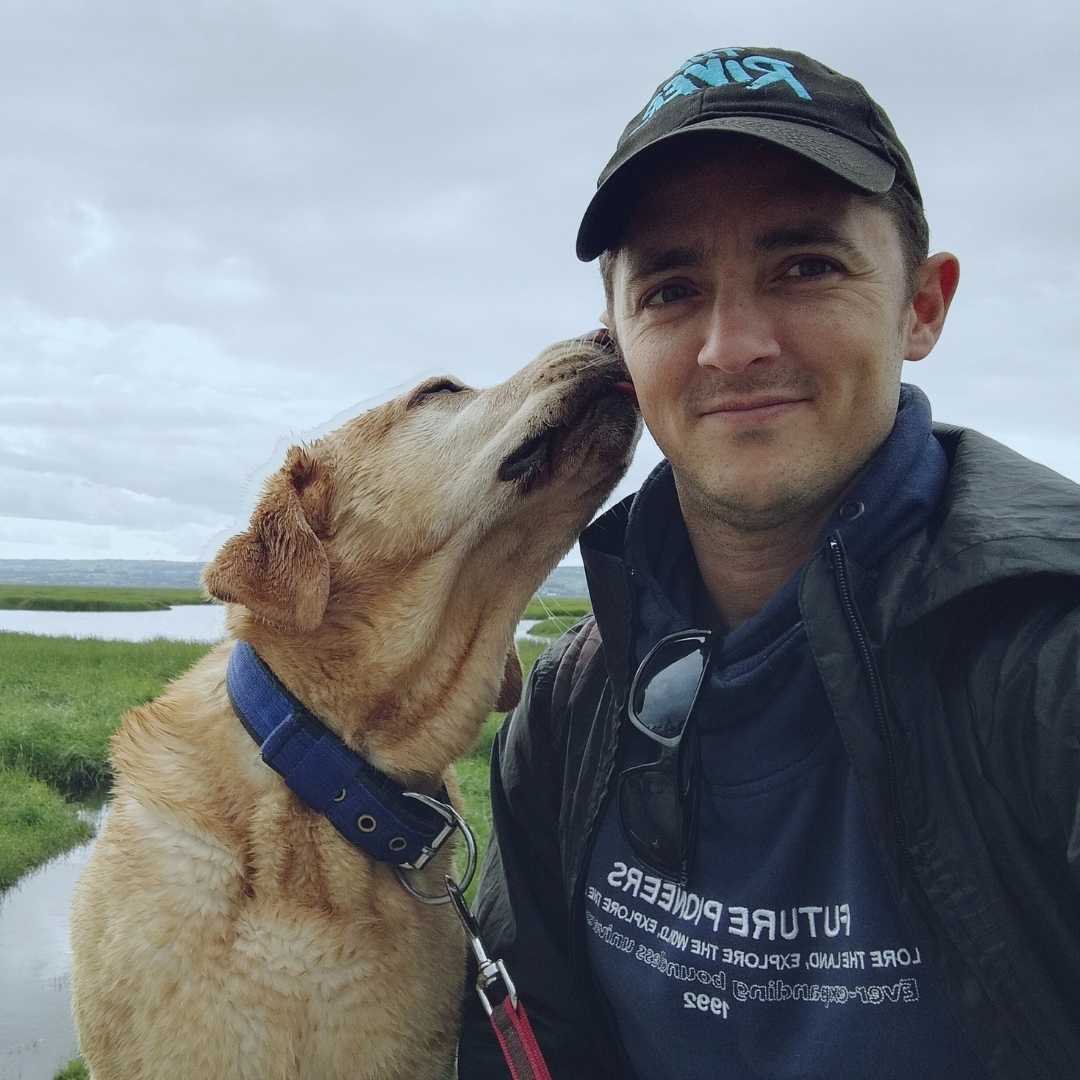 Yellow Labrador licking his owner's cheek. Owner is a white male wearing a blue jacket and baseball cap