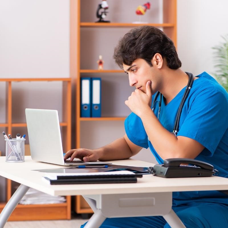 Dark haired male in blue scrubs sat as desk looking at computer in clinical setting